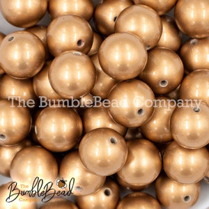 May include: A close-up image of a pile of gold-colored round beads. The beads are smooth and shiny, with a slight metallic sheen. The beads are all the same size and shape, and they are arranged in a random pattern.