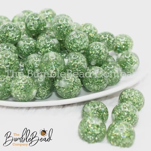 May include: A collection of green glitter beads, displayed on a white plate. The beads are round and transparent, filled with sparkling green glitter. The Bumble Bead Company logo is visible in the bottom left corner.