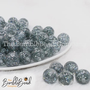 May include: A collection of clear, round beads with a glittery, iridescent interior. The beads are displayed on a white plate and scattered on a white surface. The text "The BumbleBead Company" is visible.