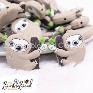 May include: Two brown silicone sloth charms with green leaves hanging from a brown branch. The sloths have white faces and black eyes. The charms are on a white fluffy surface.