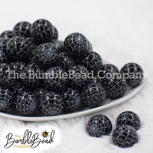 May include: A pile of black and white spiderweb patterned round beads on a white plate. The beads are arranged in a random pattern. The plate is on a white fuzzy surface.