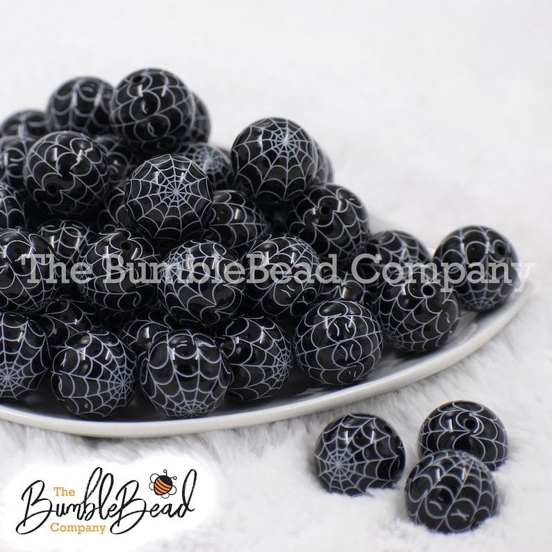 May include: A pile of black and white spiderweb patterned round beads on a white plate. The beads are arranged in a random pattern. The plate is on a white fuzzy surface.