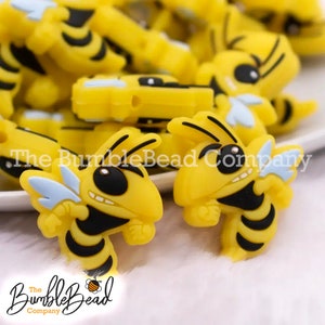 May include: A close-up of several yellow and black bee-shaped silicone charms. The bees have black eyes, black stripes, and a light blue wing. The charms are on a white surface.