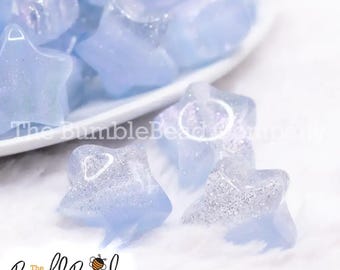 May include: A close-up of a group of light blue star-shaped resin beads with silver glitter. The beads are scattered on a white surface.