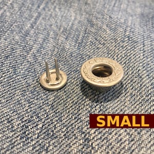 Puede incluir: Primer plano de dos botones a presión de metal plateado para jeans. El botón de la izquierda tiene dos puntas. El botón de la derecha tiene un anillo decorativo alrededor del agujero central. El texto "SMALL" es visible en la esquina inferior derecha de la imagen.
