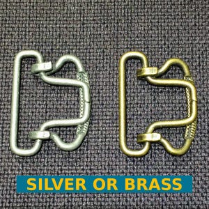 Pode incluir: Dois fechos de fivela de cinto de metal, um prateado e outro de latão. Ambos têm um design semelhante com um laço duplo.