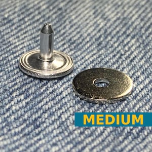Puede incluir: Un broche de presión de metal plateado con una parte superior puntiaguda y una base redonda. El broche de presión está sobre un fondo de tela vaquera azul. El texto "MEDIUM" está impreso en amarillo sobre un fondo azul.