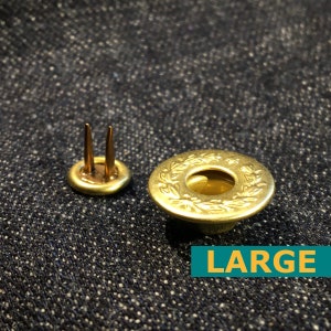 May include: Two brass-colored jean rivets. One rivet has two prongs, and the other has a floral design around the edge. The word "LARGE" is in a green box.