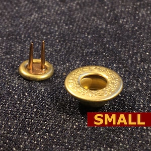 May include: A close-up of a small, gold-colored metal button with a decorative wreath design. The button is on a blue denim fabric background. The word "SMALL" is printed in red letters below the button.