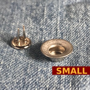 Op de afbeelding: Twee zilveren metalen jeansnieten, één met twee pinnen en de andere met een verzonken midden. Het woord "SMALL" wordt weergegeven in een rode doos. De klinknagels staan op een blauwe denim achtergrond.