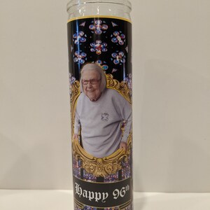 Op de afbeelding: Een glazen kaars met een zwart en gouden etiket. Het etiket toont een foto van een lachende vrouw en de tekst "Happy 96th". De achtergrond van het etiket is een glas-in-loodpatroon.