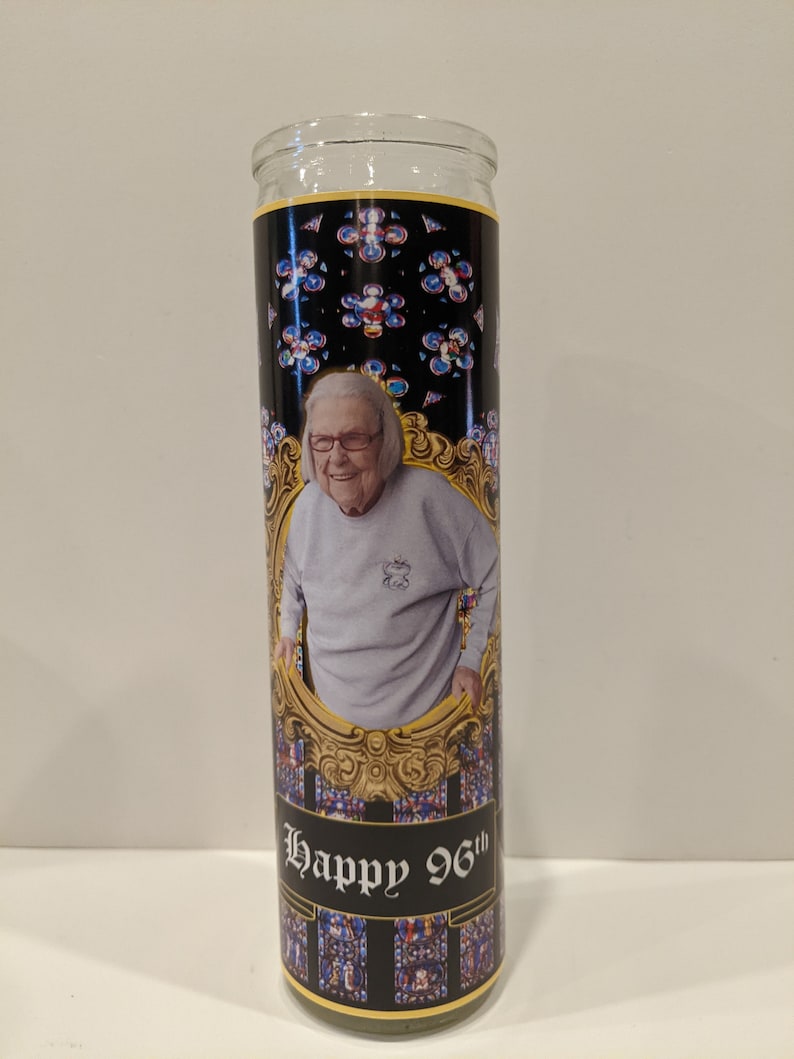 Op de afbeelding: Een glazen kaars met een zwart en gouden etiket. Het etiket toont een foto van een lachende vrouw en de tekst "Happy 96th". De achtergrond van het etiket is een glas-in-loodpatroon.