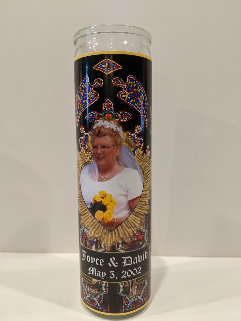 Op de afbeelding: Een witte glazen kaars met een zwart en gouden etiket. Het etiket toont een foto van een bruid die een boeket gele bloemen vasthoudt. De tekst op het etiket luidt "Joyce & David May 5, 2002".