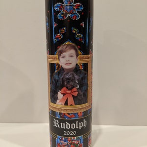 Op de afbeelding: Een zwarte en gouden kaars met een glas-in-loodontwerp. De kaars heeft een foto van een jonge jongen die een kleine zwarte hond vasthoudt. De tekst "Rudolph 2020" is op de kaars gedrukt.