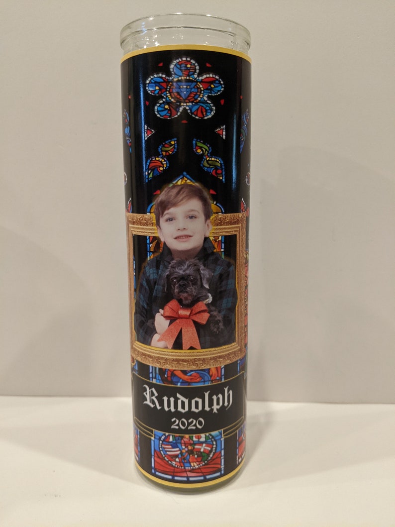 Op de afbeelding: Een zwarte en gouden kaars met een glas-in-loodontwerp. De kaars heeft een foto van een jonge jongen die een kleine zwarte hond vasthoudt. De tekst "Rudolph 2020" is op de kaars gedrukt.