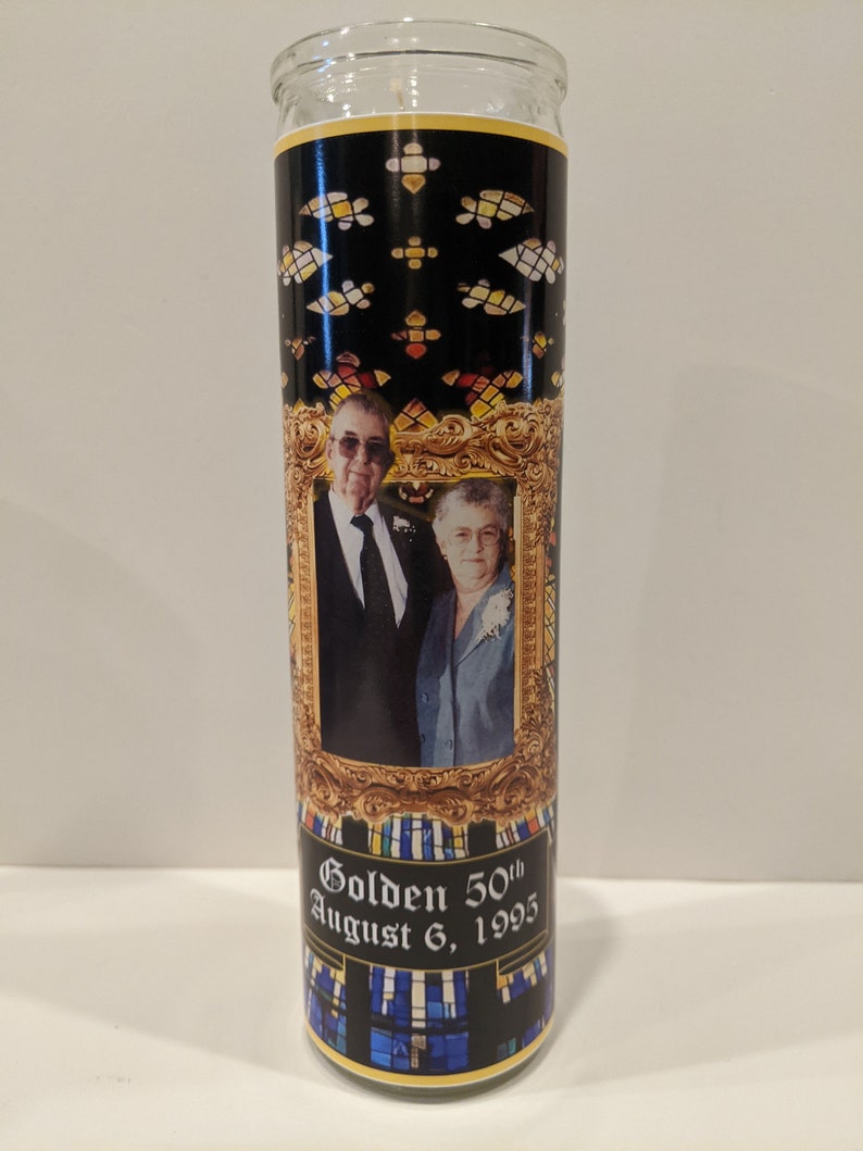 Op de afbeelding: Een glazen kaars met een zwart en gouden ontwerp. De kaars heeft een foto van een stel in een gouden lijst. De tekst "Golden 50th August 6, 1995" staat op de kaars gedrukt.
