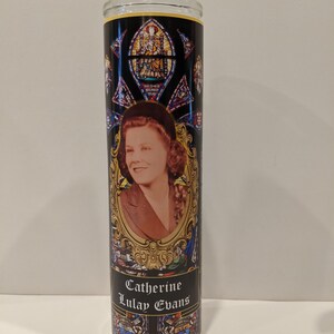 Op de afbeelding: Een zwarte en gouden glazen kaars met een glas-in-loodontwerp en een foto van een vrouw. De kaars is gelabeld "Catherine Lulay Evans".