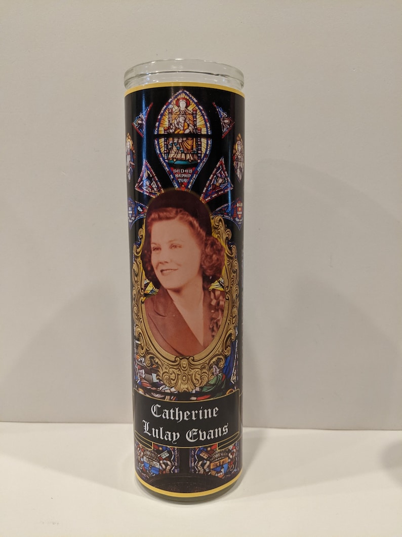 Op de afbeelding: Een zwarte en gouden glazen kaars met een glas-in-loodontwerp en een foto van een vrouw. De kaars is gelabeld "Catherine Lulay Evans".