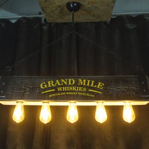 May include: A rustic wooden light fixture with the text "Grand Mile Whiskies Specialist Whisky Merchant" hanging from a chain. The fixture has five Edison-style light bulbs.