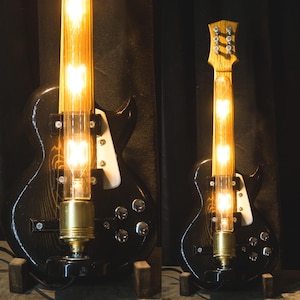 May include: Two black electric guitars with clear glass tubes containing light bulbs. The guitars are made into lamps with the light bulbs illuminating the body of the guitars.