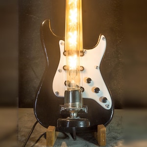 May include: A black and white electric guitar-shaped lamp with a glowing light bulb in the center. The lamp has a white pickguard, black knobs, and is supported by a wooden stand. The guitar lamp is a unique home decor item.