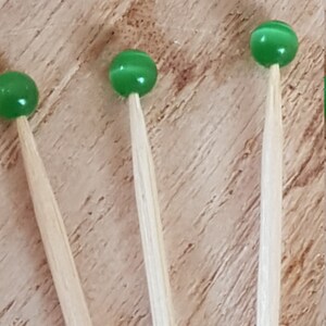 May include: Three green glass beads on wooden skewers. The beads are a light green color and have a slightly iridescent finish. The skewers are made of natural wood and are about 6 inches long.