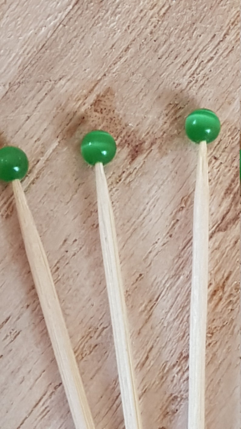 May include: Three green glass beads on wooden skewers. The beads are a light green color and have a slightly iridescent finish. The skewers are made of natural wood and are about 6 inches long.