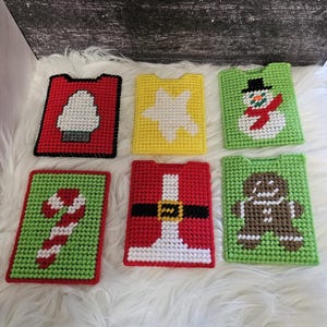 May include: Six colorful, handmade Christmas-themed plastic canvas coasters. Designs include a Christmas tree, star, snowman, candy cane, Santa suit, and gingerbread man. Each coaster is rectangular with a small tab at the top.