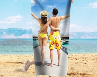 May include: A beach towel with a photo of a couple in swimsuits standing on a wooden walkway over the water. The couple is wearing yellow and floral patterned swimwear. The background is a tropical beach scene with blue water and a clear sky.