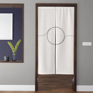 Op de afbeelding: Witte linnen kamerdeler met een zwart cirkel- en lijndesigne. De deler hangt aan een houten deurframe.