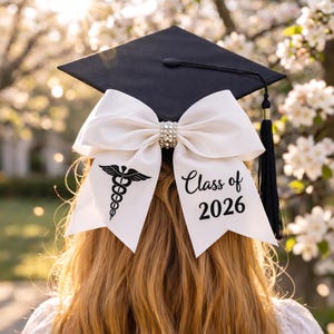 Könnte beinhalten: Eine schwarze Abschlusskappe mit einer weißen Schleife mit einem Strassstein in der Mitte. Die Schleife hat ein medizinisches Caduceus-Symbol und den Text "Class of 2026". Eine schwarze Quaste hängt von der Kappe.