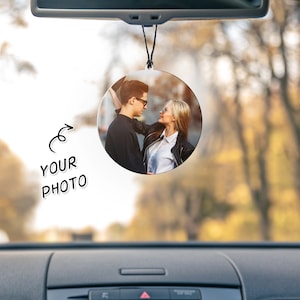Puede incluir: Un ambientador de coche redondo con una foto de una pareja mir&aacute;ndose. El ambientador est&aacute; colgado del espejo retrovisor de un coche.