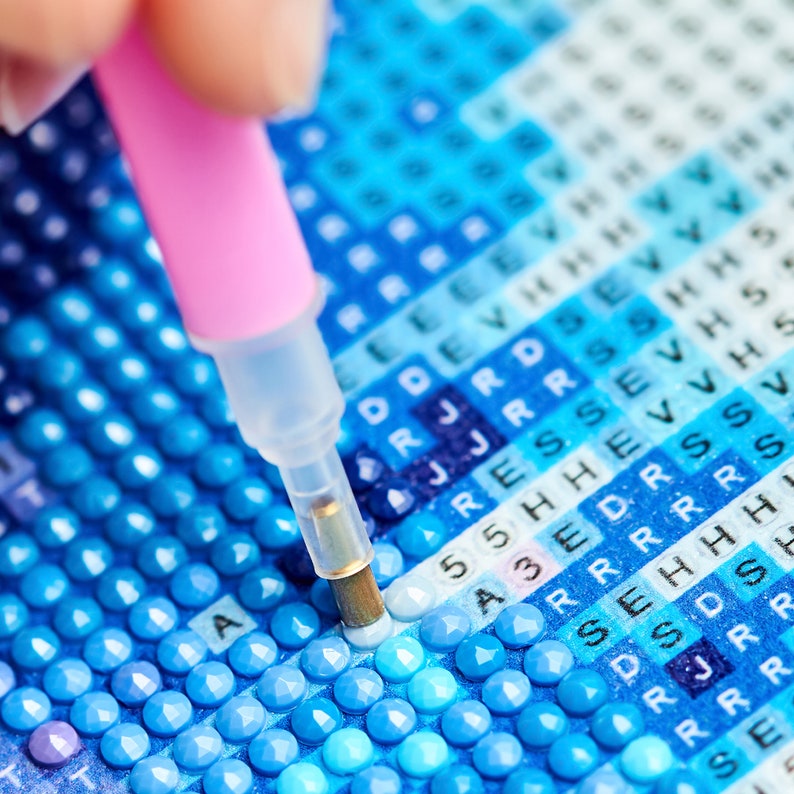 May include: Close-up of a diamond painting in progress. A pink tool is being used to place a blue diamond on the canvas. The canvas is covered in small squares with letters and numbers printed on them.