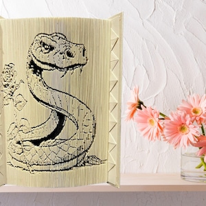 May include: A beige papercraft sculpture of a snake with a rose behind it. The snake is curled up and has a detailed, textured design.