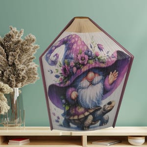 May include: A purple and white gnome wearing a hat with flowers and riding a turtle. The gnome is made from folded book pages.