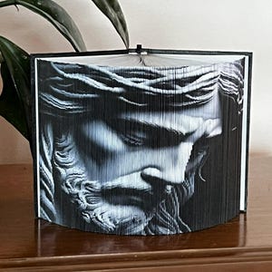 May include: A black and white book art sculpture of a man's face with a crown of thorns. The sculpture is made from folded pages of a book.