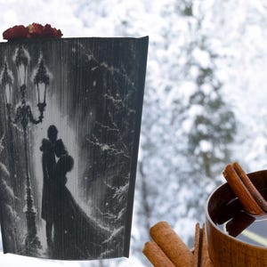May include: A black and white book sculpture showing a couple embracing under a lamppost in a snowy scene. The sculpture is topped with red flowers. A wooden mug with a warm beverage and cinnamon sticks is in the foreground.