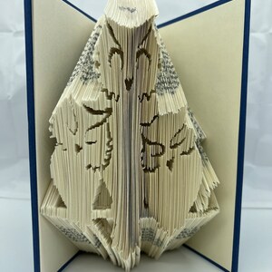 May include: A book with pages folded into a three-dimensional design of a butterfly. The book is open and the butterfly is in the center of the image. The book has a blue cover.