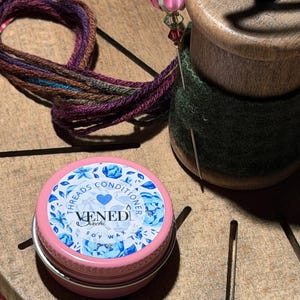 Op de afbeelding: Een roze blikje sojawas met de tekst "VENEDI" en "Threads Conditioner" in een bloemmotief. Een houten spoel met een groene vilten inzetstuk bevat een naald met een roze bloemvormige kop. Veelkleurig garen is ook zichtbaar.