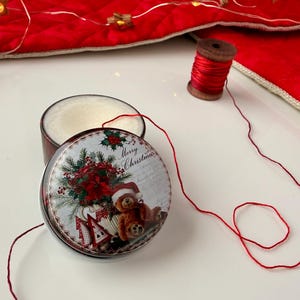 Könnte beinhalten: Eine runde, braune Dose mit einem weihnachtlichen Design, das einen Teddybären mit Weihnachtsmütze, Weihnachtssternen und den Worten "Merry Christmas" zeigt. Eine Spule roten Fadens und eine rote Schnur sind ebenfalls vorhanden.