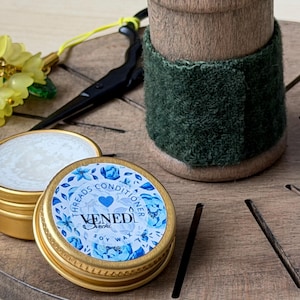 Op de afbeelding: Close-up van een goudkleurige blikje sojawas conditioner met een blauw bloemmotief en de tekst "Venedi Seche Threads Conditioner Soy Wax". Een houten spoel met een groene stoffen wikkel staat op de achtergrond. Gele bloemversieringen en een zwarte schaar zijn ook zichtbaar.