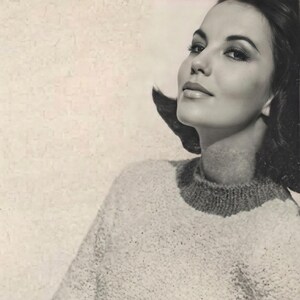 May include: A monochrome image of a woman in a textured, cream-coloured sweater. The sweater features a ribbed, dark grey collar and cuffs. The woman is leaning against a wall, with her hand on her hip.