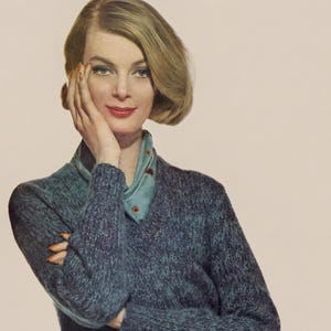 May include: A woman in a blue-grey V-neck sweater and a light blue scarf. The sweater has a textured look. She is also wearing a brown skirt. The image has a vintage aesthetic.