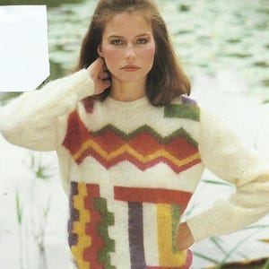 Può includere: Una donna indossa un maglione color crema con un design geometrico colorato e una gonna bianca a balze. Il maglione presenta motivi a zig-zag e rettangolari in rosso, arancione, giallo, verde e viola. La gonna ha un orlo a balze.