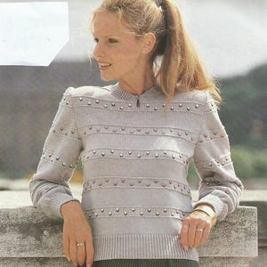 May include: A light gray sweater with horizontal stripes and small, decorative accents. The sweater has long sleeves and a round neckline with a small keyhole detail. The model is wearing gold bracelets and rings.