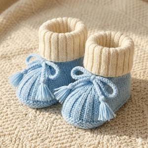 May include: A pair of knitted baby booties in light blue, with cream-coloured ribbed cuffs. Each bootie has a bow and tassel detail in matching light blue yarn. A cosy and charming design.