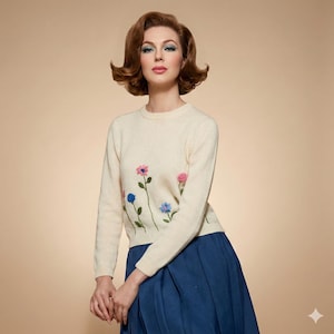 May include: A cream-coloured sweater featuring embroidered flowers in pink, blue, and purple. The sweater has long sleeves and a crew neck. The model is wearing a blue skirt. The background is a plain beige colour.