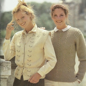 Pode incluir: Duas mulheres vestindo malhas. Uma usa um colete creme com um padrão de folhas sobre uma camisa abotoada. A outra usa um suéter de malha de cabo em uma cor marrom claro. Ambas estão sorrindo e parecem estar ao ar livre.