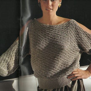 May include: A light brown, open-shoulder sweater with long sleeves. The sweater has a textured knit pattern and a ribbed hem and cuffs. The sleeves have a slit at the shoulder. The model is wearing gold earrings.
