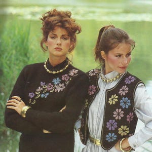 Könnte beinhalten: Zwei Frauen in schwarzen Strickwaren mit floralen Stickereien. Eine trägt einen langärmeligen Pullover, die andere eine ärmellose Weste. Beide haben goldene Halsketten und Armbänder. Der Hintergrund zeigt eine verschwommene Wasserszene. Die Weste hat einen goldenen Besatz und Gürtel.